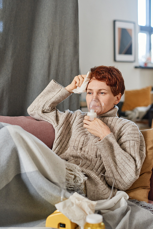 Vertical medium shot of mature Caucasian woman with short red hair having coronavirus relaxing at home using nebulizer