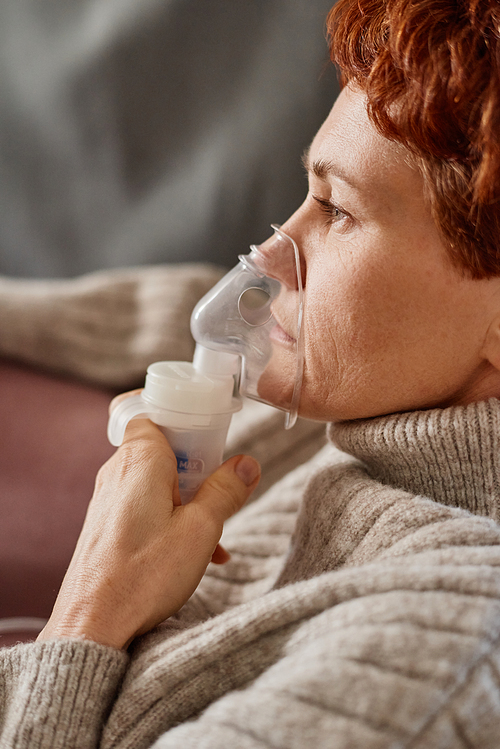 Vertical close-up side view shot of mature Caucasian woman having coronavirus relaxing at home using nebulizer