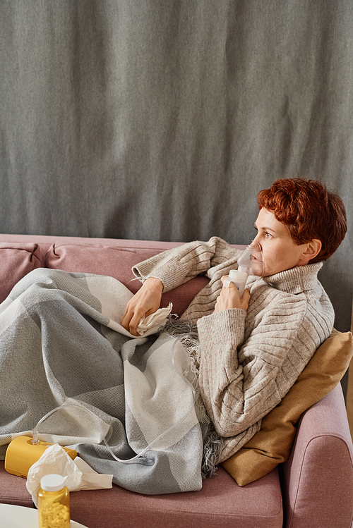 Vertical high angle shot of mature Caucasian woman suffering flu staying at home relaxing on sofa using nebulizer