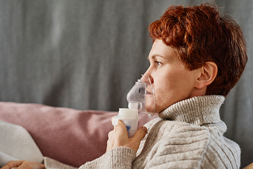 Horizontal side view close-up shot of mature woman suffering flu staying at home relaxing on sofa using nebulizer