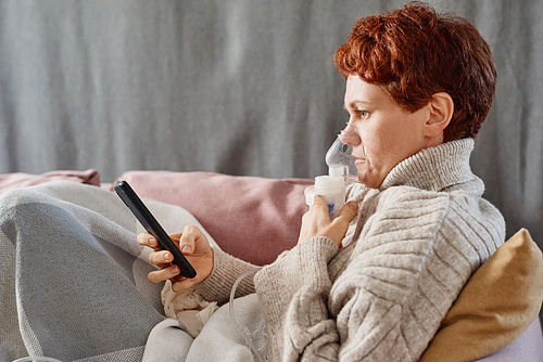 Horizontal side view shot of mature woman suffering cold staying at home relaxing on sofa using nebulizer and surfing Internet on smartphone