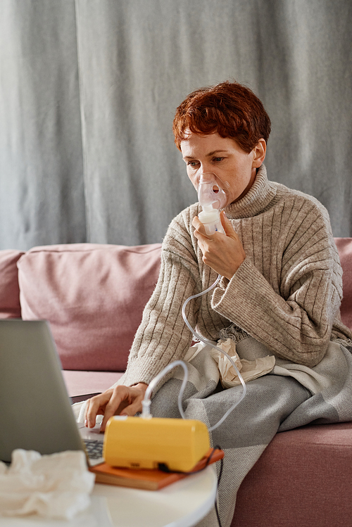 Vertical shot of mature woman suffering coronavirus staying at home sitting on sofa using nebulizer and watching film on laptop