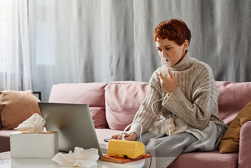 Mature woman suffering coronavirus staying at home sitting on sofa using nebulizer and surfing Internet on laptop