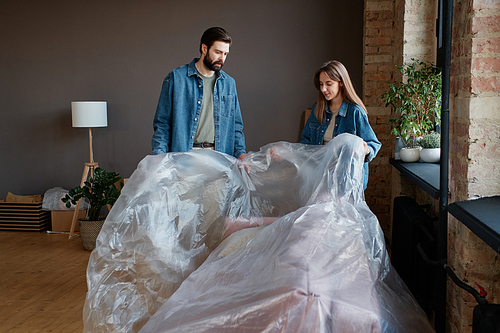 Horizontal shot of young Caucasian man and woman unpacking furniture after moving to new house taking plastic film off sofa