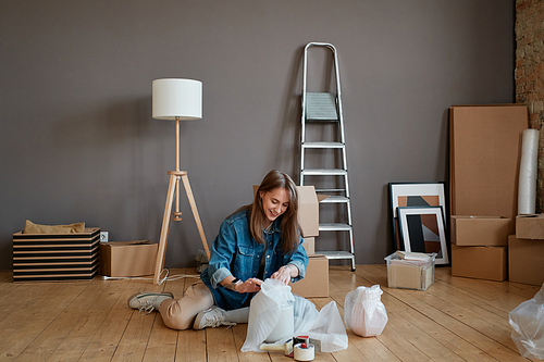 Horizontal shot of happy young Caucasian woman sitting on floor in new apartment unpacking various things after moving to it