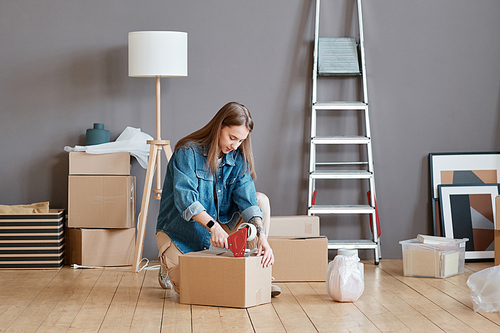 Horizontal shot of young Caucasian woman sitting on floor getting ready to move to new apartment packing things into boxes