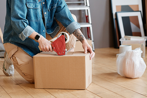 Horizontal shot of unrecognizable young woman sitting on floor getting ready for moving to new house packing things into boxes and taping them