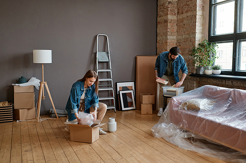 Horizontal long shot of young Caucasian man and woman moving to new apartment unpacking boxes