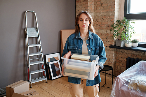 Horizontal medium portrait of young Caucasian woman moving to new apartment holding plastic box with books looking at camera