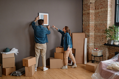 Horizontal long shot of young Caucasian man and woman choosing wall in new apartment to hang paintings on