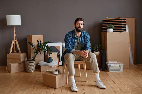 Horizontal shot portrait of young Caucasian man with beard on face sitting in loft room with stuff packed for moving to new apartment