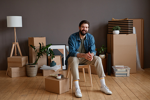 Horizontal shot portrait of young Caucasian man with beard on face sitting in loft room with stuff packed for moving to new house