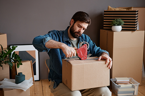 Horizontal medium shot footage of young man sitting on chair preparing for moving to new house packing things into boxes and taping them