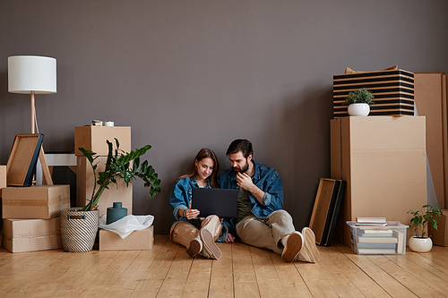 Horizontal shot footage of young man and woman sitting in new apartment surfing Internet on laptop searching for interior design pictures