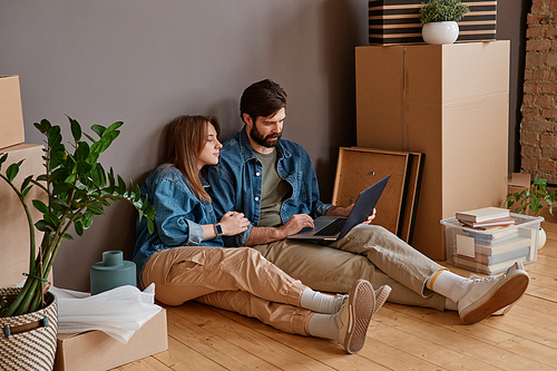 Young man and woman sitting in new apartment surfing Internet on laptop searching for interior design variants