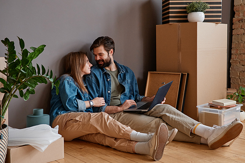 Young man and woman in love sitting on floor in new apartment surfing Internet on laptop searching for interior design variants