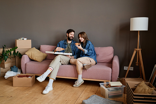 Young Caucasian man and woman relaxing together on sofa eating delicious pizza in loft living room after moving to new apartment