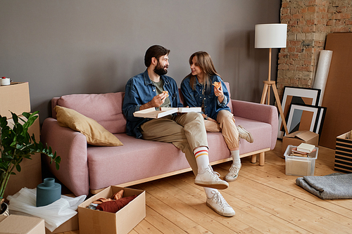 Modern young man and woman sitting together on sofa eating pizza in loft living room after moving to new apartment