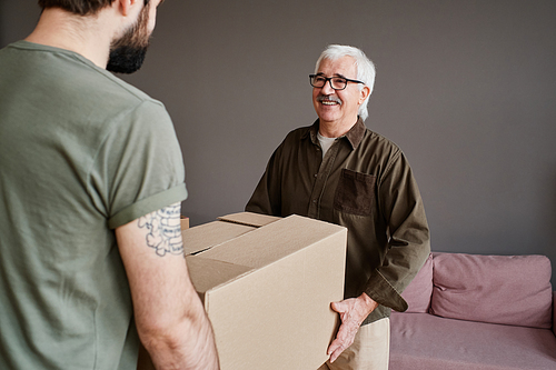 Horizontal medium shot of young man helping his senior father with moving to new house carrying big heavy box full of stuff together