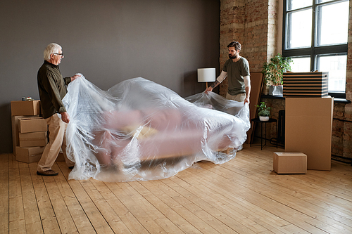 Horizontal long shot of senior Caucasian man helping his son to move to another apartment wrapping sofa with plastic film