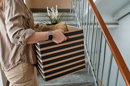 Horizontal medium section shot of unrecognizable young woman going upstairs carrying box with pillows and houseplant in it