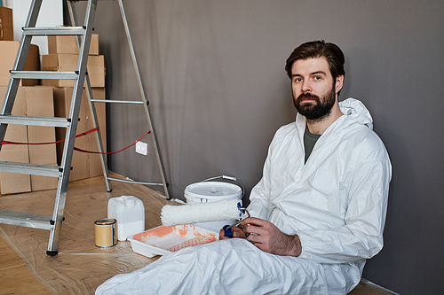 Young Caucasian man wearing protective clothes doing renovating works in new apartment sitting on floor looking at camera