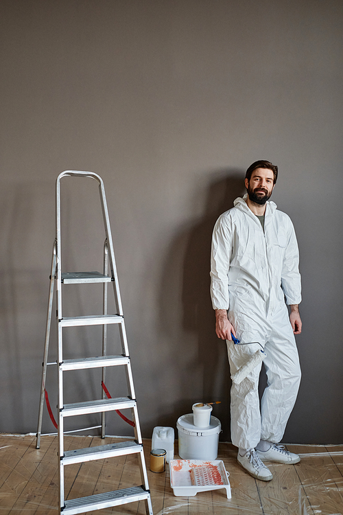 Vertical shot of handsome Caucasian man wearing protective outfit doing renovating works in his apartment standing against gray wall looking at camera