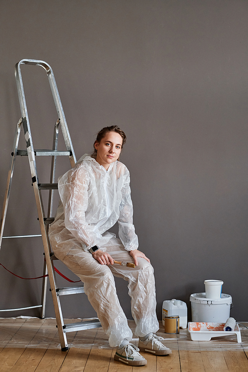 Vertical shot of beautiful young woman wearing protective clothes doing renovating works in her apartment sitting on ladder looking at camera