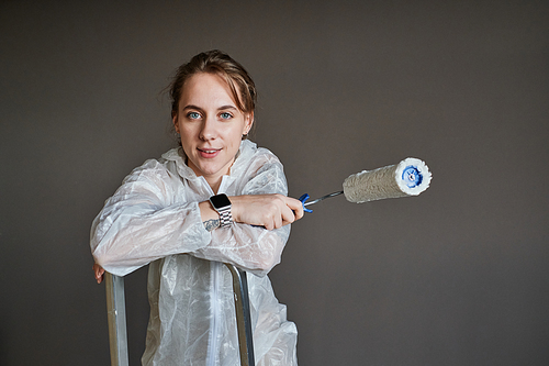 Medium portrait of beautiful young woman wearing protective clothes doing renovating works in her apartment standing on ladder against gray background