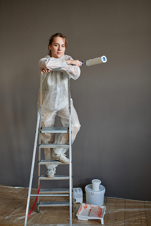 Vertical shot of attractive young woman wearing protective outfit doing repair works in her house standing on ladder looking at camera