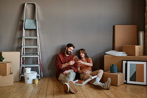 Horizontal shot of young man and woman sitting together on floor eating pizza and drinking champagne celebrating moving to loft apartment