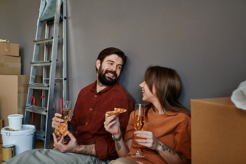 Horizontal shot of young man and woman in love sitting together on floor eating pizza and drinking sparkling wine celebrating moving to new apartment