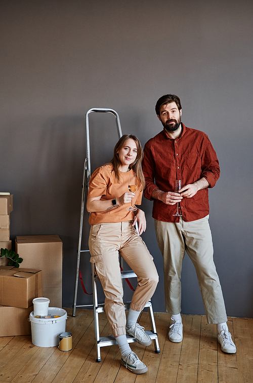 Vertical shot of young man and woman drinking sparkling wine celebrating moving to new apartment looking at camera