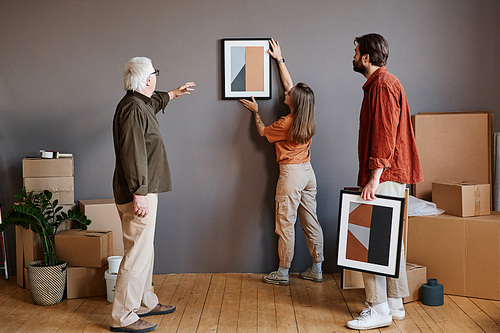 Horizontal long shot of young man and woman helping grandpa to decorate living room in new house