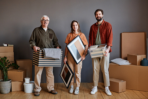 Horizontal group portrait of senior man, his son with his girlfriend standing together in loft living room in modern apartment holding various stuff